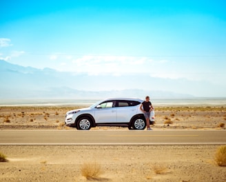 man standing beside white SUV near concrete road under blue sky at daytime