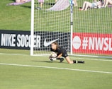 A soccer goalkeeper in a black uniform is on the ground, clutching a soccer ball near the goalpost on a field with a grassy background. Advertising boards for Nike Soccer and NWSL are visible. Spectators sit on a grassy hill behind the goal.