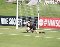 A soccer goalkeeper in a black uniform is on the ground, clutching a soccer ball near the goalpost on a field with a grassy background. Advertising boards for Nike Soccer and NWSL are visible. Spectators sit on a grassy hill behind the goal.