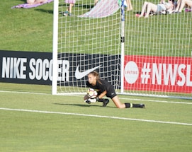 A soccer goalkeeper in a black uniform is on the ground, clutching a soccer ball near the goalpost on a field with a grassy background. Advertising boards for Nike Soccer and NWSL are visible. Spectators sit on a grassy hill behind the goal.