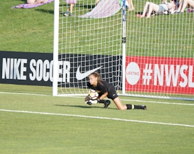 A soccer goalkeeper in a black uniform is on the ground, clutching a soccer ball near the goalpost on a field with a grassy background. Advertising boards for Nike Soccer and NWSL are visible. Spectators sit on a grassy hill behind the goal.