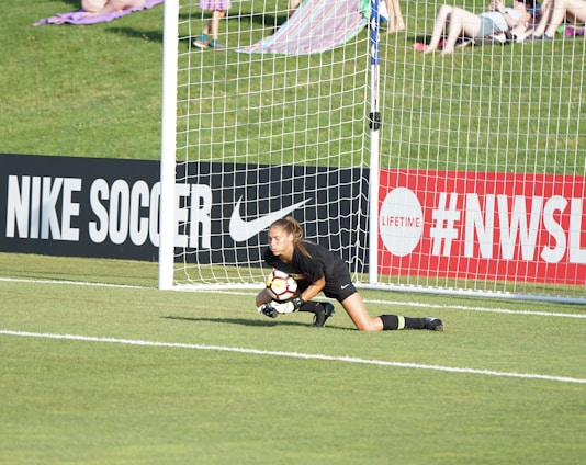 A soccer goalkeeper in a black uniform is on the ground, clutching a soccer ball near the goalpost on a field with a grassy background. Advertising boards for Nike Soccer and NWSL are visible. Spectators sit on a grassy hill behind the goal.
