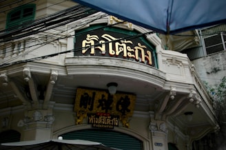 An old building with ornate architectural details features signage with Thai script in gold and black colors. The structure shows decorative trimmings and is partially obscured by an umbrella in the foreground. Above, numerous black cables are visible against the overcast sky.