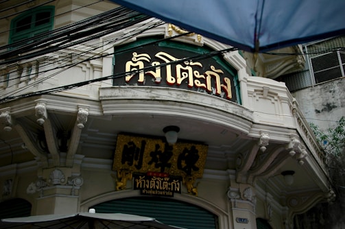 An old building with ornate architectural details features signage with Thai script in gold and black colors. The structure shows decorative trimmings and is partially obscured by an umbrella in the foreground. Above, numerous black cables are visible against the overcast sky.