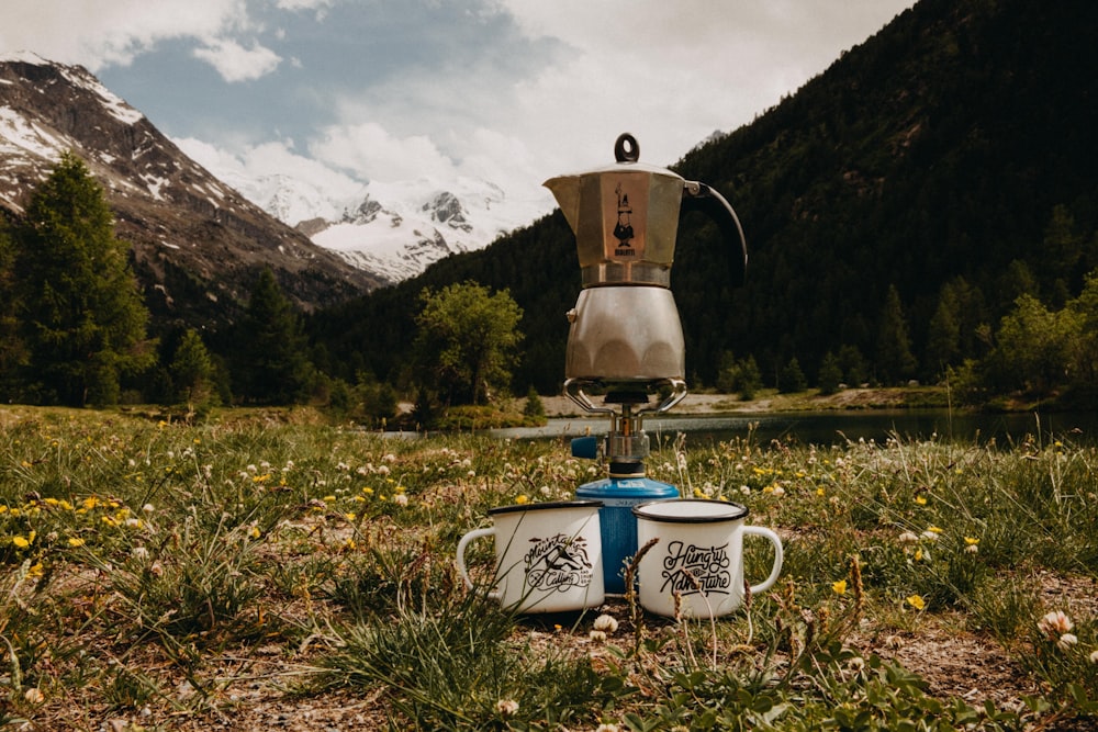 grauer Edelstahl-Mokapot auf dem Brenner zwischen zwei weißen Bechern auf grünem Gras