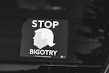 A black and white sticker is displayed on a surface, featuring the silhouette of a hairstyle resembling a well-known figure. The text 'STOP BIGOTRY' is prominently visible, conveying a political message. The sticker includes a reference to the year 2016 and a website, DCCC.org.