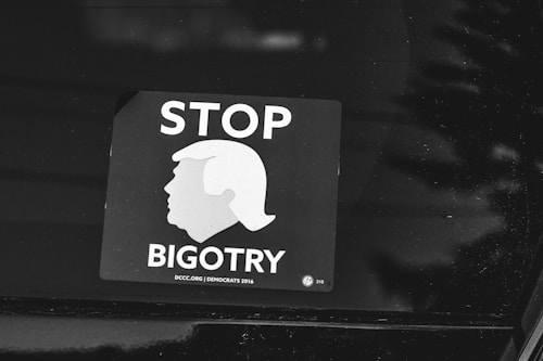 A black and white sticker is displayed on a surface, featuring the silhouette of a hairstyle resembling a well-known figure. The text 'STOP BIGOTRY' is prominently visible, conveying a political message. The sticker includes a reference to the year 2016 and a website, DCCC.org.