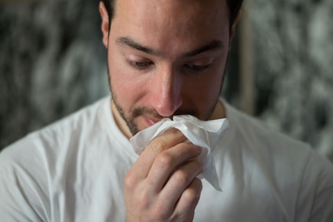 A man holds a tissue to his nose, appearing contemplative or in the midst of a cold. He wears a white shirt and looks downwards.