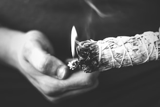 A monochrome image showing a person holding a bundle of smudge sticks, likely sage, which is being lit by a lighter held in the other hand. Smoke can be seen rising, suggesting the ritual or ceremonial use of the smudge sticks. The image focuses closely on the hands and the smudge stick with a blurred background.