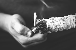 A monochrome image showing a person holding a bundle of smudge sticks, likely sage, which is being lit by a lighter held in the other hand. Smoke can be seen rising, suggesting the ritual or ceremonial use of the smudge sticks. The image focuses closely on the hands and the smudge stick with a blurred background.