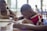 A group of children are seated at a table, intently working on writing or drawing with paper and pens. The environment appears to be well-lit with natural light coming through the windows in the background.
