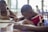 A group of children are seated at a table, intently working on writing or drawing with paper and pens. The environment appears to be well-lit with natural light coming through the windows in the background.