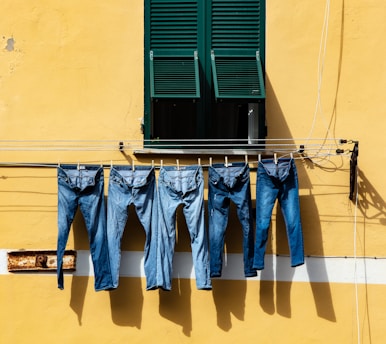 A row of five pairs of blue jeans hang on a clothesline against a bright yellow wall. The line is positioned beneath a green window with shutters partially open. The wall has a horizontal white stripe and some visible wear.