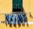 A row of five pairs of blue jeans hang on a clothesline against a bright yellow wall. The line is positioned beneath a green window with shutters partially open. The wall has a horizontal white stripe and some visible wear.