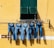 A row of five pairs of blue jeans hang on a clothesline against a bright yellow wall. The line is positioned beneath a green window with shutters partially open. The wall has a horizontal white stripe and some visible wear.