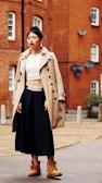 A person stands confidently in an urban setting, wearing a beige trench coat over a white crop top, navy blue wide-leg pants, and brown boots. The background features a red brick building with multiple satellite dishes and windows.