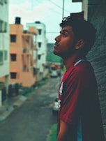 A man gazes upwards with a contemplative expression, standing beside a building. The background features a blurry urban street with residential buildings and parked vehicles.