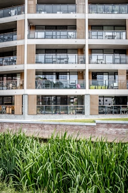 A modern multi-story apartment building features large glass balconies and wood paneling accents. The architecture is minimalistic and contemporary, with clean lines and a focus on symmetry. In the foreground, tall green grass grows alongside a brick wall, providing a natural element to the urban environment.