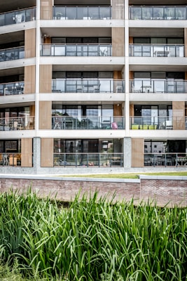 A modern multi-story apartment building features large glass balconies and wood paneling accents. The architecture is minimalistic and contemporary, with clean lines and a focus on symmetry. In the foreground, tall green grass grows alongside a brick wall, providing a natural element to the urban environment.