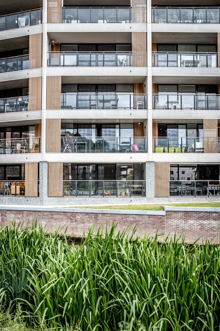 A modern multi-story apartment building features large glass balconies and wood paneling accents. The architecture is minimalistic and contemporary, with clean lines and a focus on symmetry. In the foreground, tall green grass grows alongside a brick wall, providing a natural element to the urban environment.