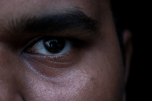 A close-up shot focuses on the left eye of a person, highlighting skin texture and eyebrow details. The lighting creates subtle shadows and reflections in the eye.