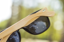 A close-up view of sunglasses with wooden frames and dark lenses. The lenses reflect a serene sky with a few clouds and silhouettes of trees. The background is softly blurred, featuring a mix of green and yellow shades.