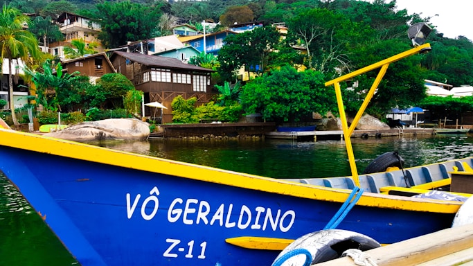 A vibrant scene with a blue and yellow boat named 'Vô Geraldino Z-11' is docked in front of lush greenery and colorful hillside homes. The houses are nestled among trees, and the water reflects their colors, creating a lively and picturesque view.