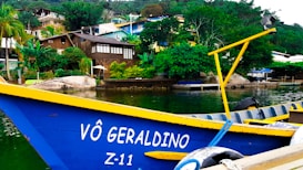 A vibrant scene with a blue and yellow boat named 'V&ocirc; Geraldino Z-11' is docked in front of lush greenery and colorful hillside homes. The houses are nestled among trees, and the water reflects their colors, creating a lively and picturesque view.