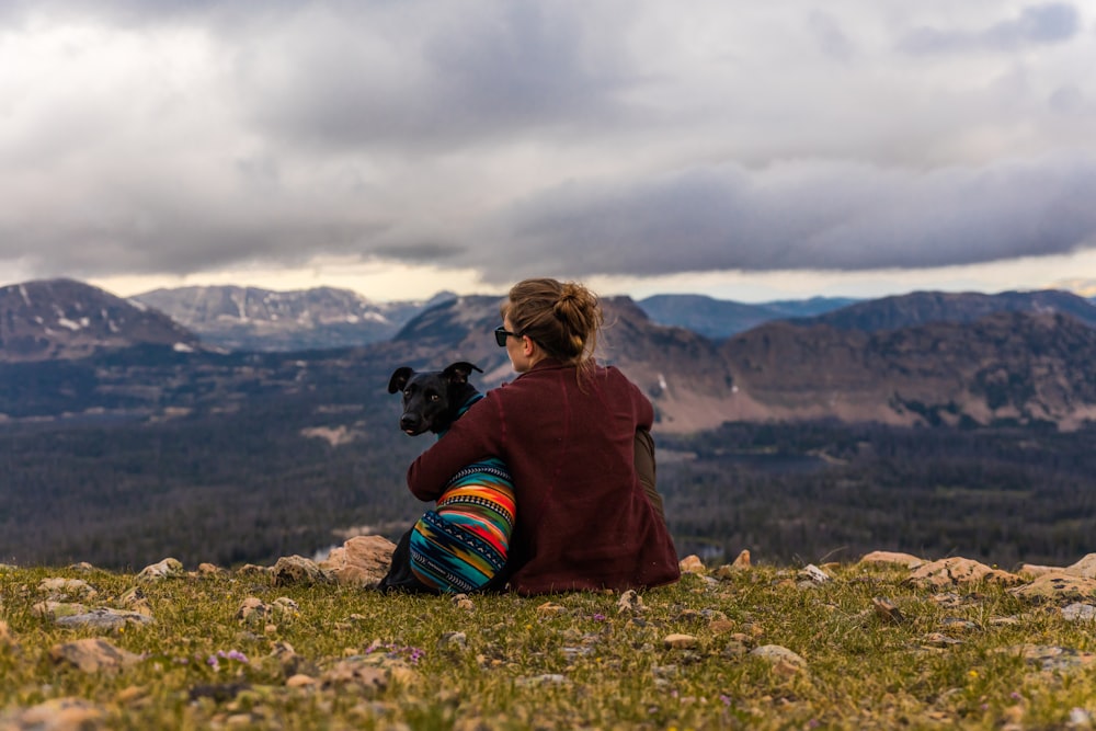 woman sitting near dog on cliff overlooking mountains and forest during daytime
