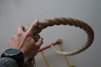 A hand gripping a thick rope, with a watch on the wrist, and a blurred background that includes gym equipment.