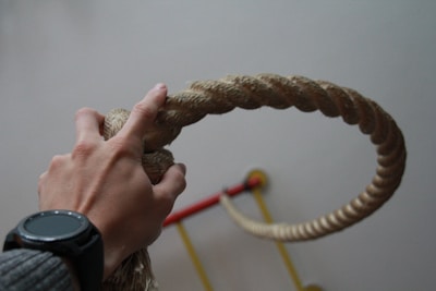 A hand gripping a thick rope, with a watch on the wrist, and a blurred background that includes gym equipment.