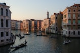 Gondolas glide along a serene canal flanked by historic, colorful buildings under a warm, setting sun. The architecture features classic arches and detailed façades, creating a picturesque cityscape. A bell tower rises in the background, adding to the charming ambiance.