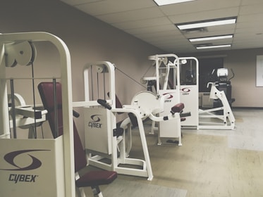 A gym room featuring several Cybex exercise machines arranged neatly. The machines are predominantly white with red seating, and the room has a clean, orderly appearance with overhead fluorescent lighting. The walls are a neutral color, and the flooring appears to be a mix of beige and light wooden tones.