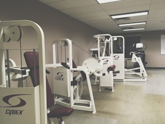 A gym room featuring several Cybex exercise machines arranged neatly. The machines are predominantly white with red seating, and the room has a clean, orderly appearance with overhead fluorescent lighting. The walls are a neutral color, and the flooring appears to be a mix of beige and light wooden tones.