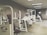 A gym room featuring several Cybex exercise machines arranged neatly. The machines are predominantly white with red seating, and the room has a clean, orderly appearance with overhead fluorescent lighting. The walls are a neutral color, and the flooring appears to be a mix of beige and light wooden tones.