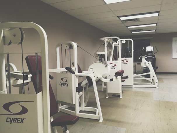 A gym room featuring several Cybex exercise machines arranged neatly. The machines are predominantly white with red seating, and the room has a clean, orderly appearance with overhead fluorescent lighting. The walls are a neutral color, and the flooring appears to be a mix of beige and light wooden tones.