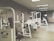 A gym room featuring several Cybex exercise machines arranged neatly. The machines are predominantly white with red seating, and the room has a clean, orderly appearance with overhead fluorescent lighting. The walls are a neutral color, and the flooring appears to be a mix of beige and light wooden tones.