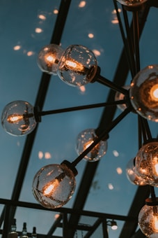 A ceiling light fixture featuring multiple round glass bulbs with visible filaments, suspended from a black metal framework. The background shows the structure of a roof with a soft blue sky visible through it.