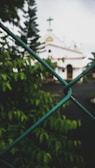 A view through a chain-link fence with a blurred background showing a white church building with a cross on top. The foreground features green foliage in focus, while the sky appears overcast.