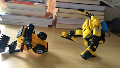 Two yellow and black toy figures made of interlocking bricks are placed on a wooden surface. One appears to be a robot with a humanoid form, showing articulation in its limbs, while the other resembles a construction vehicle, possibly a bulldozer or excavator, tipped over on its side. In the background, stacks of books are visible.