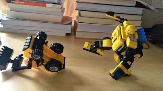 Two yellow and black toy figures made of interlocking bricks are placed on a wooden surface. One appears to be a robot with a humanoid form, showing articulation in its limbs, while the other resembles a construction vehicle, possibly a bulldozer or excavator, tipped over on its side. In the background, stacks of books are visible.