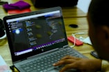 A person is working on a laptop displaying code in a text editor. The environment is a wooden table with various items including a pair of headphones, a red external hard drive, and some documents. The screen shows lines of code, indicating software development or programming activity.