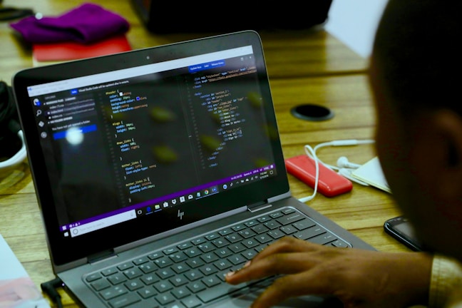 A person is working on a laptop displaying code in a text editor. The environment is a wooden table with various items including a pair of headphones, a red external hard drive, and some documents. The screen shows lines of code, indicating software development or programming activity.