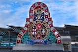 A vibrant, ornate sign featuring multiple circular emblems and symbols, with a prominent '1864' at the top. The sign is adorned with red, white, and blue colors, with decorative elements and the words 'London Chatham and Dover Railway' in gold. The background shows a modern architectural structure with a glass facade and metallic beams beneath a partly cloudy sky.