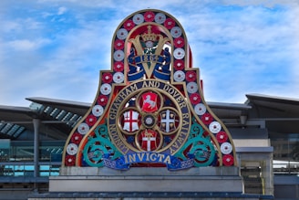 A vibrant, ornate sign featuring multiple circular emblems and symbols, with a prominent '1864' at the top. The sign is adorned with red, white, and blue colors, with decorative elements and the words 'London Chatham and Dover Railway' in gold. The background shows a modern architectural structure with a glass facade and metallic beams beneath a partly cloudy sky.