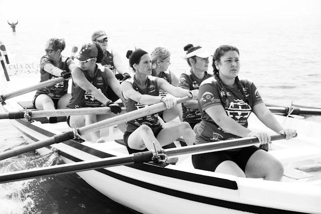 A group of women are rowing on a boat in a coordinated effort. They are all wearing matching jerseys and seem to be focused on their task. The background is the open sea, and it appears to be a cloudy day.