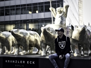 A row of bronze bear statues is positioned in front of a modern glass building. Among the bears, a person in casual attire, wearing a hat and a Raiders sweatshirt, is sitting with a relaxed pose. The statues appear strong and majestic, while the background of the building reflects a modern urban setting.