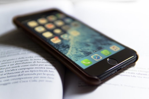 A smartphone with a visible home screen is lying on an open book. The phone is in focus, displaying app icons, while the book text is slightly blurred. The setting appears to be well-lit with natural light.