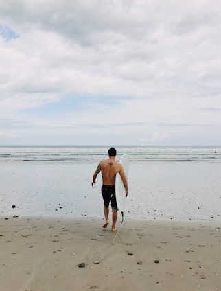 A person with a tattoo on their back is walking towards the ocean while holding a surfboard. The beach has scattered small rocks and the sky is cloudy with hints of blue.