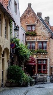 A quaint narrow street lined with old brick buildings, including a charming establishment with red-framed windows and a sign reading 'Vlaamsche Pot.' The façade is adorned with greenery and flowers. The cobblestone path and cozy outdoor seating evoke a sense of vintage European architecture.